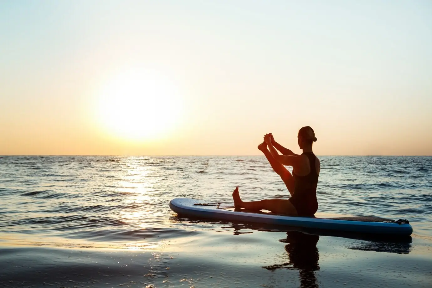 Silhouette of a beautiful woman performing yoga on a surfboard at sunrise.